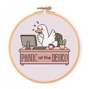May include: A cross-stitch embroidery in a round wooden hoop featuring a white goose with an orange beak sitting at a pink desk. The goose looks stressed with blue sweat drops. On the desk are a computer, a mug, and a plant. Text reads PANIC at the DESKO.