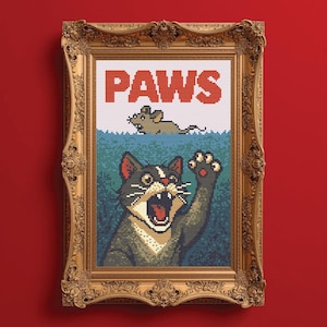 May include: Cross-stitch art in a gold ornate frame. The artwork features a cat with an open mouth and raised paw, with the word "PAWS" above a mouse in water. The background is a red wall.