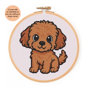 May include: A cross-stitch embroidery hoop featuring a brown cartoon dog with large, expressive eyes. The dog is stitched on a light pink fabric. The wooden hoop has a metal clasp. Text on the image says, "Comes with an alphabet so you can add your dog's name!"