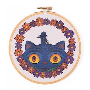 May include: Cross-stitch embroidery of a blue cat wearing a witch hat, surrounded by a floral wreath in an embroidery hoop. The cat has large orange eyes and is centered in the design. The flowers are orange and purple.