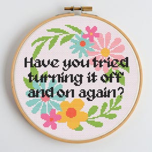 Peut inclure: Un cercle de broderie au point de croix avec le texte "Have you tried turning it off and on again ?" entouré de fleurs colorées et de feuilles vertes. Les fleurs sont roses, jaunes et bleues.