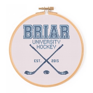 Könnte beinhalten: Ein Kreuzstich-Stickrahmen mit dem Text "BRIAR UNIVERSITY HOCKEY" in Blau. Zwei gekreuzte Hockeyschläger und ein Puck befinden sich unter dem Text. Der Text "EST. 2015" ist ebenfalls enthalten. Der Rahmen ist aus Holz.