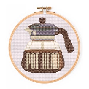 May include: Cross-stitch embroidery in a wooden hoop. The design features a coffee pot with the words "POT HEAD" stitched in brown. The coffee pot has a purple lid and handle, with a clear glass body.