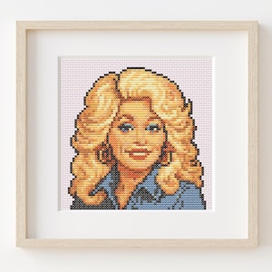May include: Framed cross-stitch portrait of a woman with blonde hair, blue eyes, and a denim shirt. The artwork is displayed in a light wood frame with a white mat, set against a pink background.