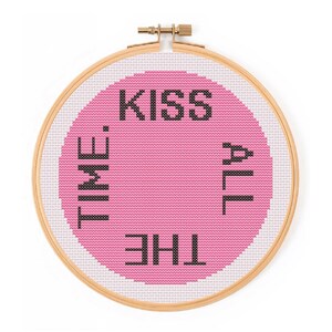 May include: A circular wooden embroidery hoop containing white Aida fabric with a large pink circle stitched in the center. The black cross-stitch text reads KISS ALL THE TIME in a circular pattern around the pink shape, with the words oriented to be read clockwise.