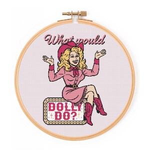 Puede incluir: Bastidor de bordado de punto de cruz con una ilustración rosa de una mujer con un atuendo rosa, botas de vaquero y sombrero de vaquero. El texto "What would DOLLY DO?" se muestra encima de la mujer y en un letrero. El bastidor es de madera.