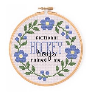 Könnte beinhalten: Ein Kreuzstich-Stickrahmen mit dem Text "Fictional Hockey Boys ruined me". Das Design zeigt blaue Blumen und grüne Blätter um den Text herum. Der Rahmen ist aus Holz und hat einen Messingverschluss.
