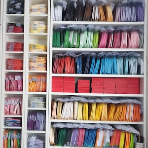 May include: A white shelf unit with multiple shelves filled with stacks of colorful fabric squares. The fabric squares are arranged in rows and columns, creating a visually appealing pattern.