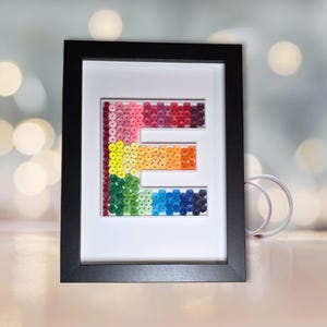 Può includere: Un'opera d'arte incorniciata con la lettera "E" realizzata con perline colorate. Le perline sono disposte in un gradiente di colori, dal rosa al blu, all'interno di un passe-partout bianco e una cornice nera. L'opera è esposta su una superficie chiara.