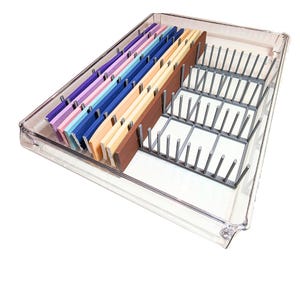 Op de afbeelding: Een doorzichtige plastic lade-organizer met meerdere compartimenten. De organizer bevat kleurrijke verdelers in tinten paars, roze, blauw en bruin. Er is ook een grijs metalen rek aanwezig, ontworpen om items rechtop te houden. De organizer is ontworpen voor gebruik in de keuken of op kantoor.