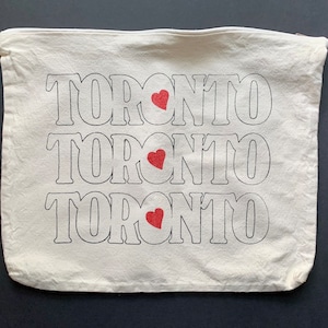 May include: A white canvas tote bag with the word "Toronto" printed three times in black, with a red heart in between each word.