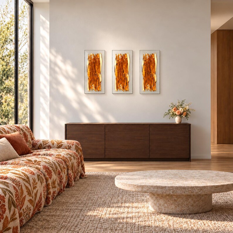 Three amber fused glass wall art panels hang in a row above a dark wooden cabinet with a small floral arrangement, beside a patterned sofa in a sunlit living room.