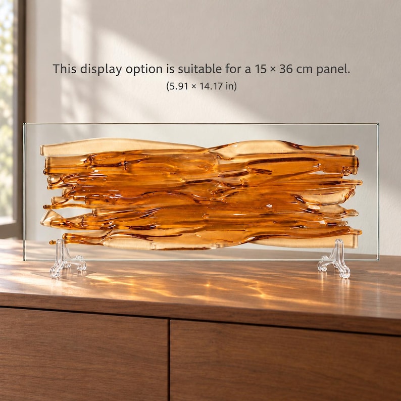Amber fused glass wall art displayed horizontally on two clear easels on a wooden cabinet, glowing in sunlight; the caption identifies this display option for a 15 x 36 cm panel.