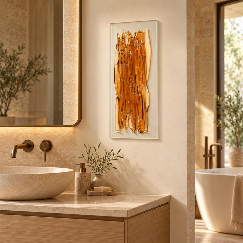 Amber fused glass wall art beside a large mirror in a warm neutral bathroom, above a stone counter and wooden vanity, with sunlight illuminating its sculptural texture.