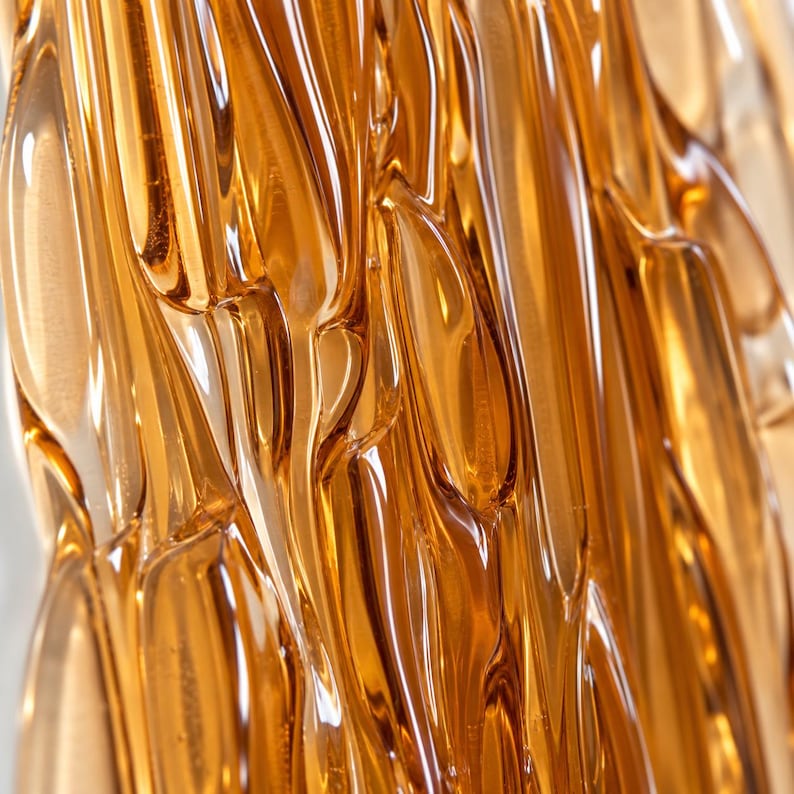Close-up of amber fused glass wall art, revealing layered ribbon-like glass, flowing ridges and clear edges that catch the light in warm honey tones.