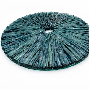 Puede incluir: Una obra de arte circular con un agujero central. Está hecha de vidrio texturizado y en capas en tonos turquesa y verde azulado. El vidrio tiene un aspecto rugoso y natural, y la pieza descansa sobre una base de vidrio transparente. Diámetro aproximado de 30 cm.