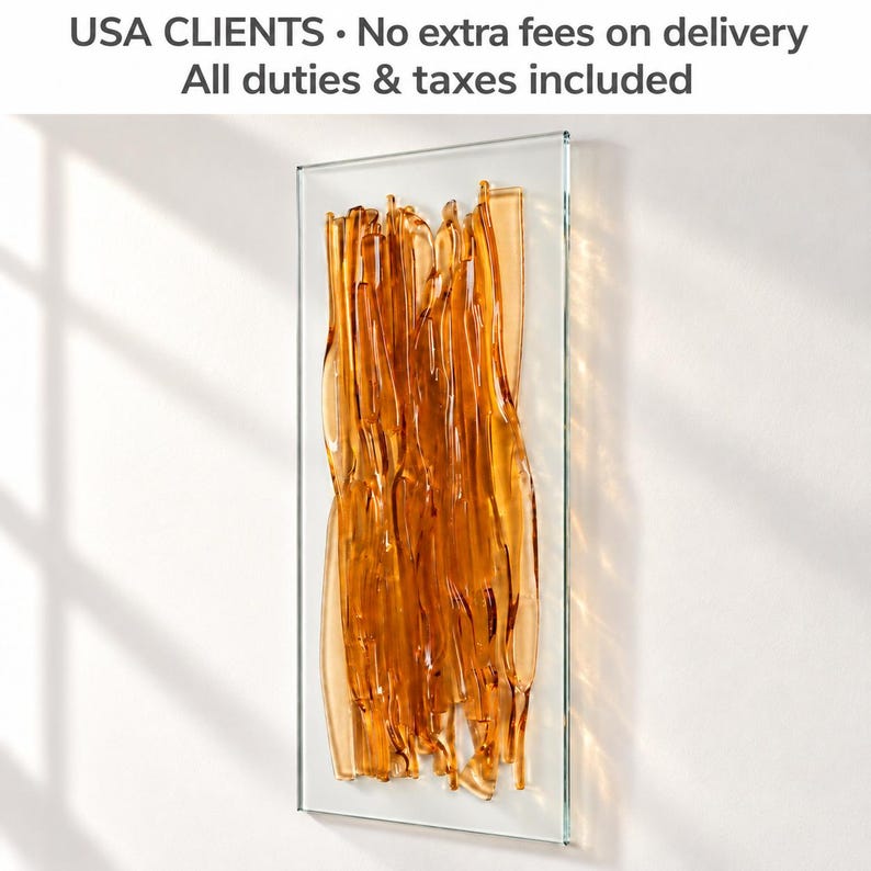 Amber fused glass wall art on a sunlit white wall, with delicate reflections and a banner stating that USA clients pay no extra delivery fees, with duties and taxes included.