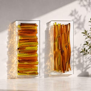 May include: Two rectangular glass wall art pieces. One features stacked amber and yellow glass pieces, the other has vertical amber glass strips. Both are encased in clear glass frames, creating a modern, abstract design. The art is displayed on a white wall.