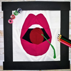 Puede incluir: Una obra de arte textil que representa labios rosas estilizados con una cereza roja y un tallo verde. La obra está enmarcada en negro y rodeada de materiales de costura y lápices de colores.