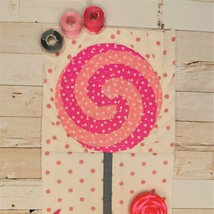 Op de afbeelding: Een roze en wit gestippelde lolly quiltblok met een grijze stok. Het blok is omgeven door spoelen garen en een roze en witte lolly.