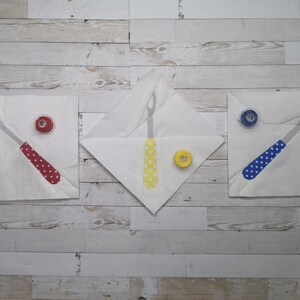 May include: Three white fabric squares with a sewing tool and spool of thread on each. The tools are red, yellow, and blue with white polka dots. The spools of thread are red, yellow, and blue.