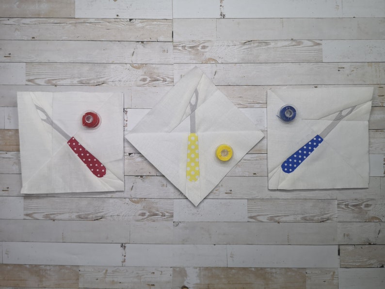 May include: Three white fabric squares with a sewing tool and spool of thread on each. The tools are red, yellow, and blue with white polka dots. The spools of thread are red, yellow, and blue.