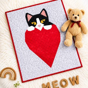 Könnte beinhalten: Handgefertigte Steppdecke mit einer schwarz-weißen Katze, die aus einem großen roten Herzen hervorschaut. Die Steppdecke hat einen grauen Hintergrund und einen roten Rand. Ein Teddybär und Holzbuchstaben mit der Aufschrift "MEOW" liegen daneben.