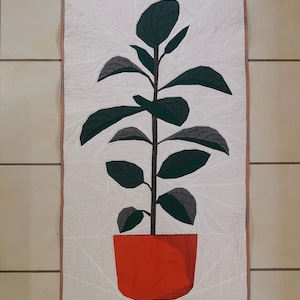 May include: A white quilted wall hanging featuring a green plant in a red pot. The plant has large, green leaves and a brown stem.