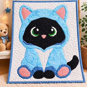 Puede incluir: Un tapiz de pared acolchado con un gato negro con un atuendo con capucha azul claro. El gato tiene grandes ojos verdes, orejas rosas y una nariz rosa. El fondo es blanco con un patrón de remolino y el edredón está bordeado de azul.