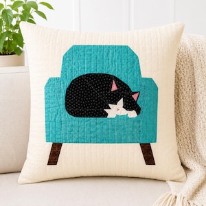 May include: A cream-coloured square cushion with a quilted pattern. It features a teal armchair design with a black and white cat sleeping inside. The cat has pink ears and a white chest. The chair has brown legs.