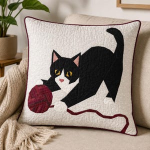 Puede incluir: Un cojín cuadrado con un diseño acolchado de un gato blanco y negro jugando con un ovillo de lana rojo oscuro. El gato tiene ojos amarillos y orejas rosas sobre un fondo blanco texturizado con un borde ribeteado en rojo oscuro.
