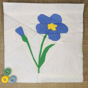 Puede incluir: Un cuadrado de tela blanca con una flor azul y un tallo verde aplicados sobre él. La flor tiene un centro amarillo.