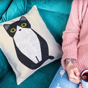 May include: Square cushion featuring a black and white cat illustration with yellow eyes. The cushion is placed on a teal sofa. A person is holding a mug of mulled wine. The cushion is likely 40cm x 40cm.