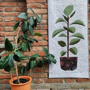 May include: A potted rubber plant with large, glossy green leaves stands beside a quilted wall hanging. The wall hanging depicts a rubber plant in a patterned pot. The plant is in an orange pot. The brick wall provides a rustic backdrop.