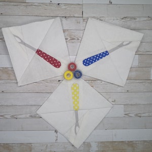 May include: Three white fabric squares with a red, blue, and yellow sewing tool design. The tools are all the same shape and have white polka dots. The squares are arranged in a triangle shape with the points facing inward. Three spools of thread are in the center of the triangle, one red, one blue, and one yellow.