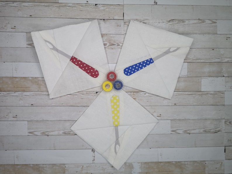 May include: Three white fabric squares with a red, blue, and yellow sewing tool design. The tools are all the same shape and have white polka dots. The squares are arranged in a triangle shape with the points facing inward. Three spools of thread are in the center of the triangle, one red, one blue, and one yellow.