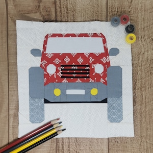 May include: A fabric block featuring a red and gray car design. The car has white headlights and a black grill. The background is white with a geometric pattern. Colored pencils and spools of thread are also visible.