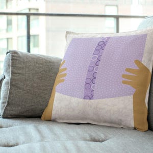 May include: Square decorative pillow with a light purple book design. The pillow has a white background with a floral pattern and tan-colored hands holding the book. The pillow is on a gray couch.