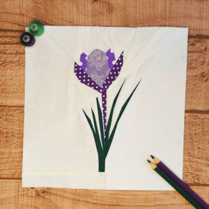 May include: A purple and white fabric flower with green leaves, created using the paper piecing technique. The flower is on a white background.