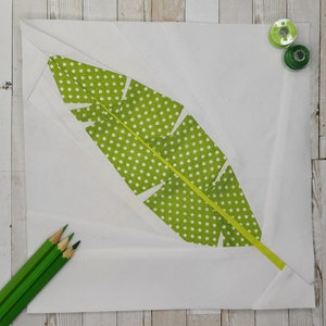 Puede incluir: Un bloque de edredón de tela cuadrado que presenta una forma de hoja verde central con lunares blancos sobre un fondo blanco sólido. La hoja tiene una vena central de color amarillo brillante. Tres lápices verdes están abajo a la izquierda y dos carretes de hilo arriba a la derecha.