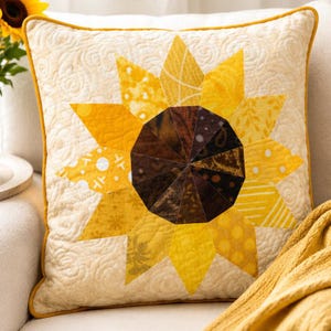 Könnte beinhalten: Quadratisches Zierkissen mit Sonnenblumenmuster. Das Kissen hat einen gesteppten cremefarbenen Hintergrund mit einer gelben Sonnenblume in der Mitte. Die Sonnenblume hat eine braune Mitte und gelbe Blütenblätter mit verschiedenen Mustern. Das Kissen hat einen gelben Rand.