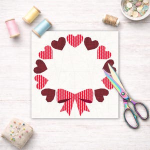 Op de afbeelding: Een stoffen hartkrans met een rood-witte ruitstrik en afwisselend effen rode en ruitjes harten. De krans staat op een witte achtergrond met naaispullen, waaronder een schaar en klosjes garen.