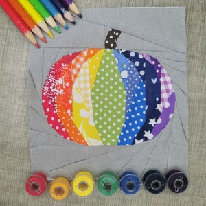 Peut inclure: Un bloc de patchwork fait main avec un motif de citrouille arc-en-ciel. La citrouille est composée de tissus aux motifs variés dans les tons de rouge, orange, jaune, vert, bleu et violet. Des crayons de couleur et des bobines de fil assorties sont disposés autour du bloc.