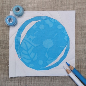 Op de afbeelding: Een wit vierkant van stof met een blauwe cirkel in het midden. De cirkel heeft een witte omtrek en een blauw bloemenpatroon. Twee spoelen blauwe draad bevinden zich in de linkerbovenhoek van de afbeelding.