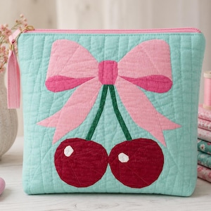 May include: A quilted mint green zippered pouch featuring a large pink bow centered above two dark red cherries with green stems. The pouch has a matching pink fabric zipper pull and a textured, stitched surface pattern throughout the fabric.
