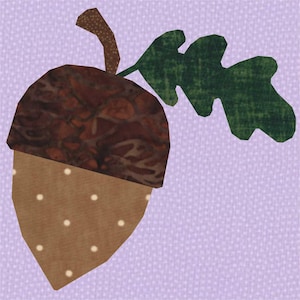 May include: A fabric acorn applique with a brown cap and tan body dotted with white circles. A green leaf and brown stem are attached to the acorn, set against a lavender background. This design is suitable for fall-themed crafts.