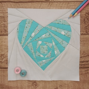 Pode incluir: Um bloco de patchwork em forma de coração feito à mão. O coração é feito de tecido turquesa com estampa floral, rodeado por tecido cinza. Dois lápis de cor e duas bobinas de linha no fundo. Sobre uma superfície de madeira.