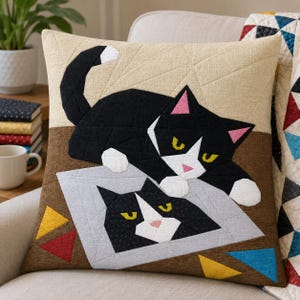 Può includere: Un cuscino quadrato con un design trapuntato raffigurante due gatti bianchi e neri. Il gatto in alto è sdraiato con la coda curva, mentre quello in basso è raffigurato come un volto all'interno di un quadrato grigio chiaro. Lo sfondo è beige e marrone.