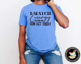 May include: A light blue t-shirt with the text "I MATCH ENERGY SO HOW WE GON' ACT TODAY" in black script. The shirt is paired with blue jeans. A round logo is visible on the bottom right.