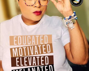 May include: A white t-shirt with the text "EDUCATED MOTIVATED ELEVATED MELANATED" printed in black, brown, and beige. The text is stacked vertically, with each word on a separate line. The shirt is being worn by a person with short, dark hair and glasses.